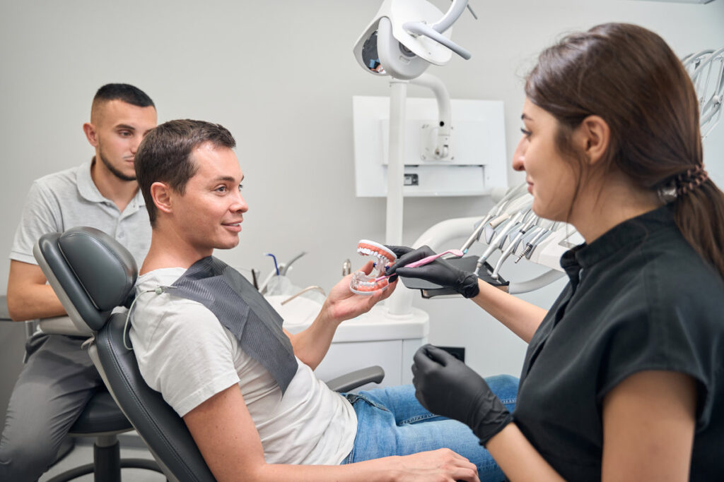 Qualified woman dentist holding 3d jaw model to explain way of dental implants installation to male client who sitting in dental chair, male doctor assisting