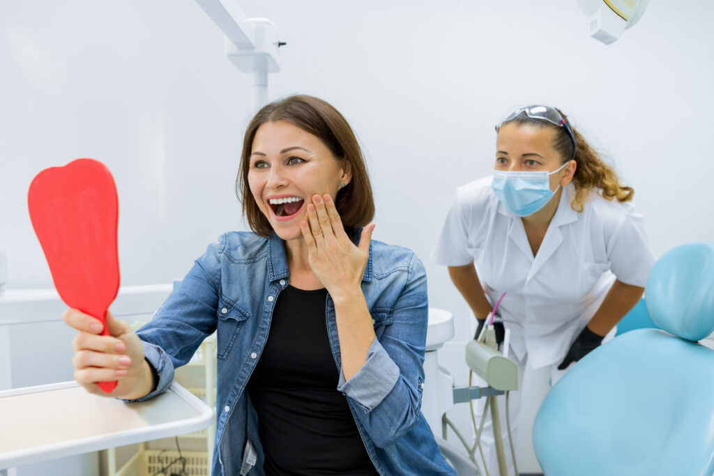 Happy mature woman after treatment and prosthetics looking in the mirror at teeth while sitting in dental chair