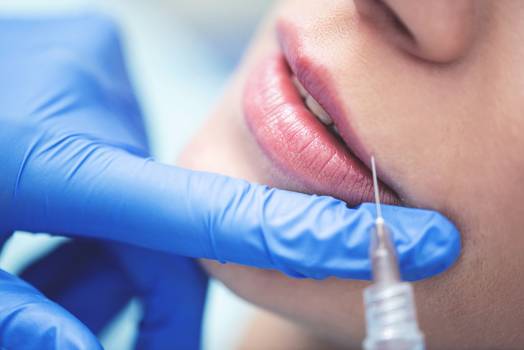 Close up of hands of cosmetologist making injection in female lips. She is holding syringe. The young beautiful woman is receiving procedure with enjoyment. Cropped picture