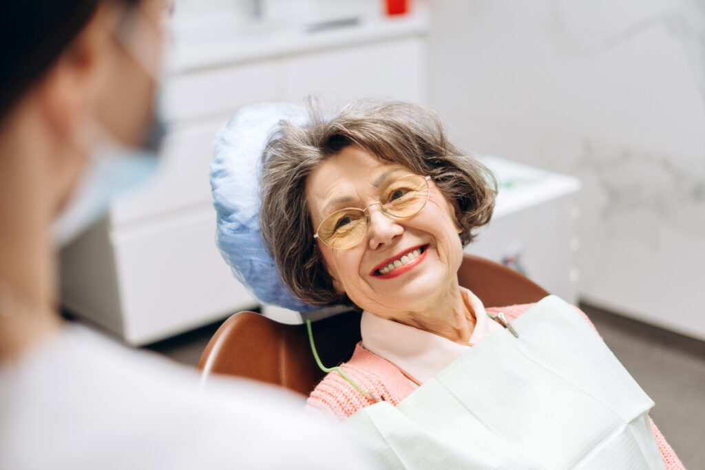 Elderly woman with glasses is smiling while sitting in a dentist chair, wearing a bib and headrest cover, suggesting a positive experience during her dental checkup. Health care concept