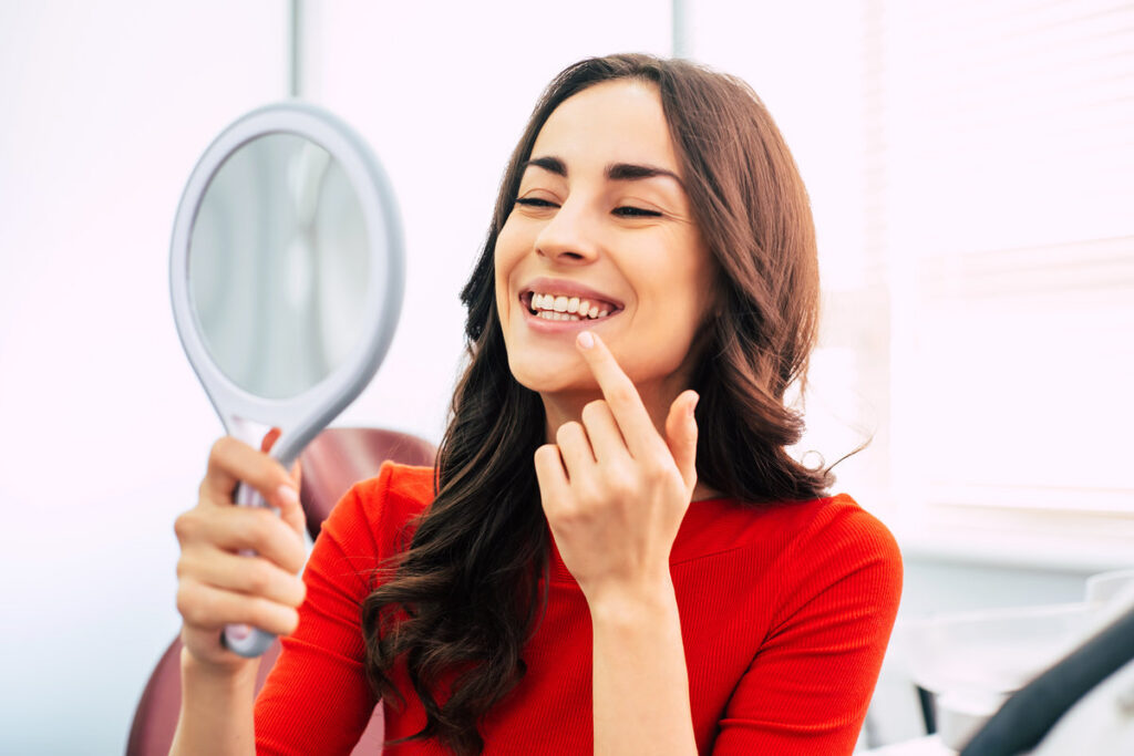 Tooth range. Hilarious girl with long curly hair in the dental chair of tooth center is using a mirror to see the result of the stomatology work which was done perfectly.