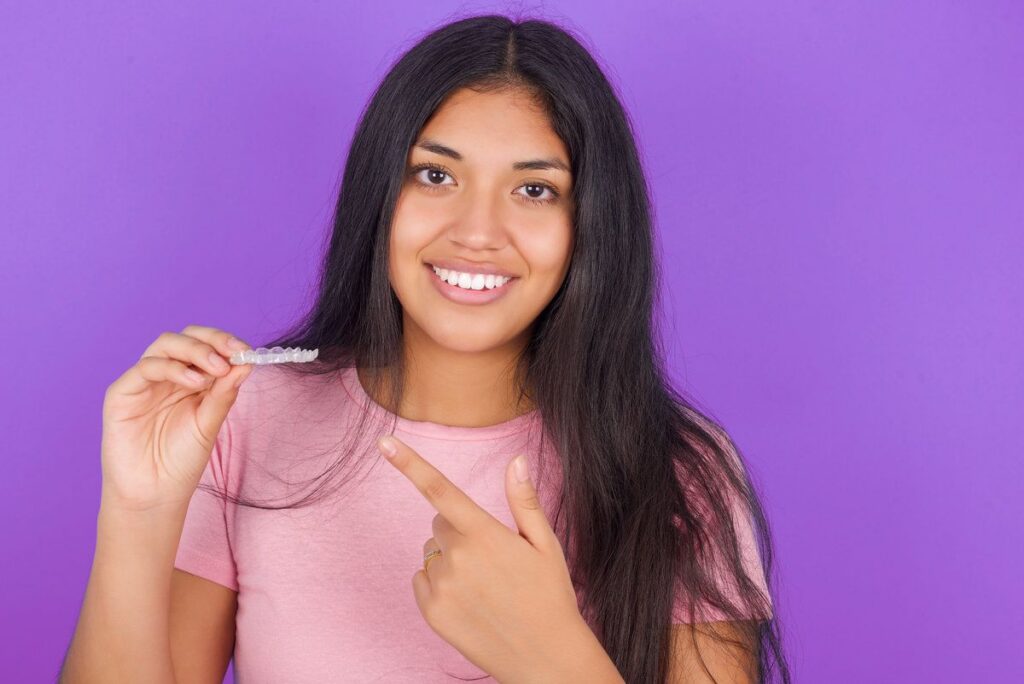 Hispanic brunette girl wearing pink t-shirt over purple background holding an invisible aligner and pointing at it. Dental healthcare and confidence concept.